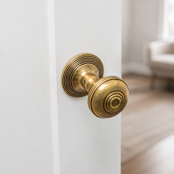 An antique brass doorknob and matching backplate mounted on a white door, with a softly blurred interior room in the background.